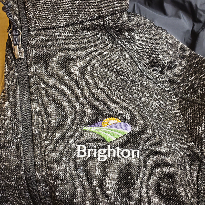 Close-up of a dark gray corporate jacket with colorful embroidered Brighton logo, showcasing professional custom embroidery by Zamar Printing